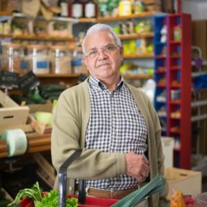Elderly man shopping in a grocery store with a basket of fresh vegetables.